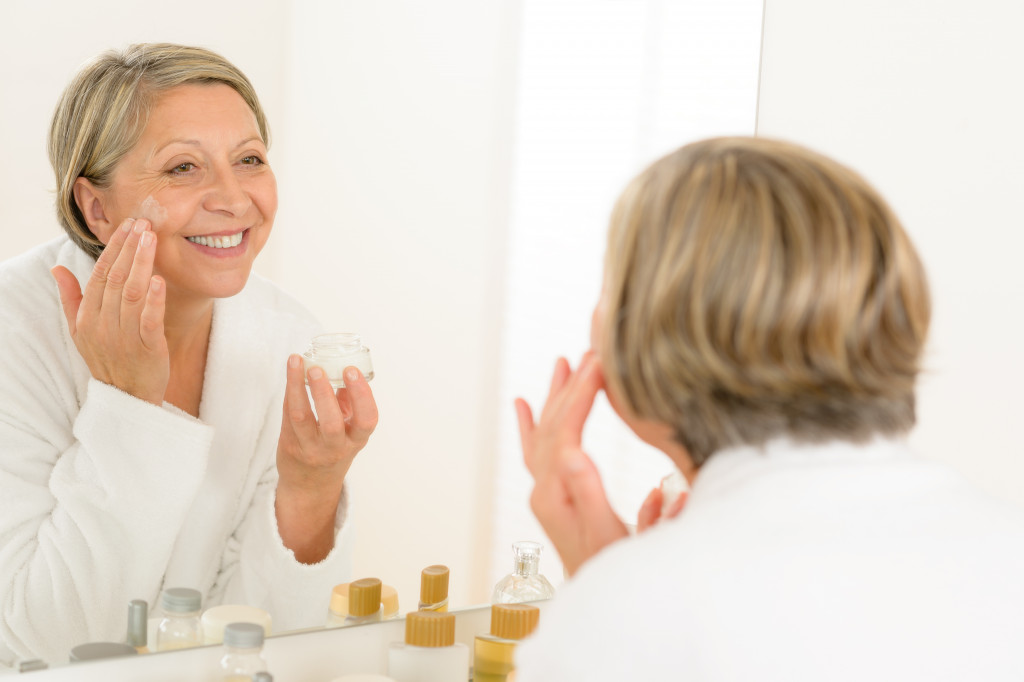 smiling elderly woman applying cream to face while looking at mirror