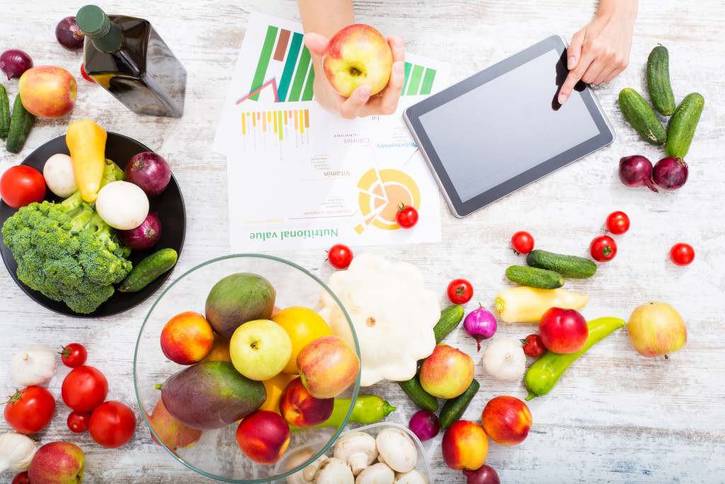 person holding apple while touching tablet in table with lots of fruits and veggies