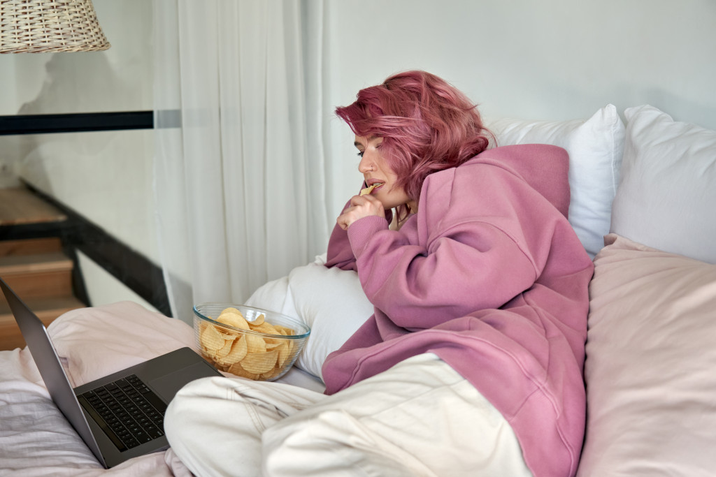 woman using laptop wating unhealthy snack in bed